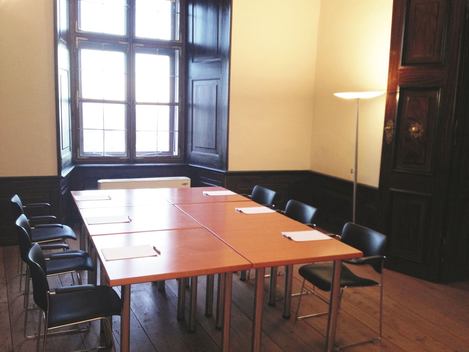 Seminarraum Schloss5a mit zusammengestellten Tischen und Sesseln. Im Eck beleuchtet eine Stehlampe den Raum
