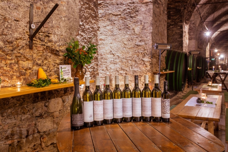 Im Bischöflichen Weinkeller ist auf einem Holztisch das Weinsortiment von Schloss Seggau schön aufgereiht. Im Hintergrund sieht man die alten Mauern und Holzfässer des Bischöflichen Weinkellers