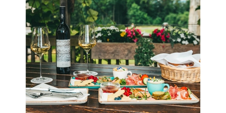 Ein vollgedeckter Tisch im Garten mit einer Flasche Wein und zwei Gläsern. Davor zwei Schmankerlteller mit regionalen Köstlichkeiten (Wurst, Speck, Käse, Marmelade u.v.m.) und ein Brotkorb