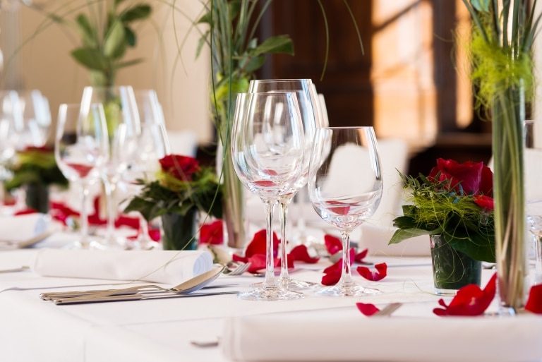 A beautifully laid table with shiny wine glasses and red flowers on the white tablecloth