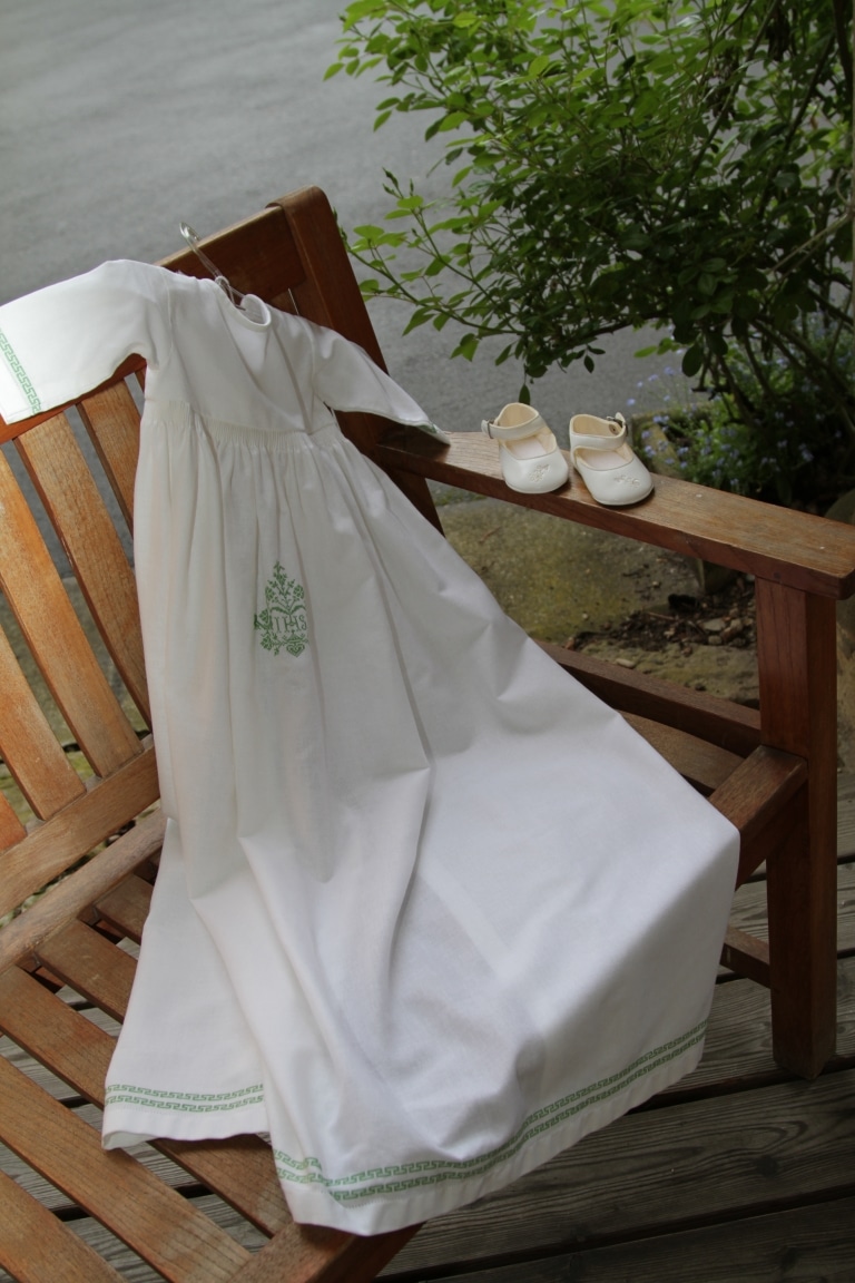 A white baptims dress is lying on a chair. Small schoes are standing on the armchair back