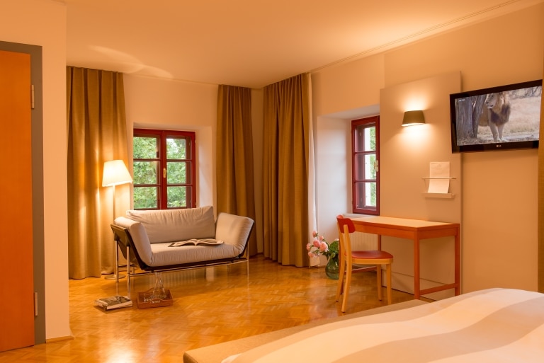 Comfort double room of Schloss Seggau with a big double bed, a cosy reading corner, a small desk an a great view over the green surrounding